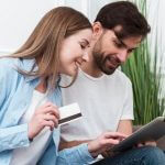 Mulher segurando um cartão e rindo, ao lado de um homem com um tablet, ambos sorrindo, representando o tema Como usar o cartão de crédito.