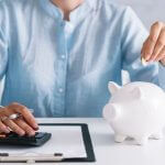 Mulher colocando dinheiro no cofre ao lado de uma calculadora, ilustrando o tema como começar a poupar'.