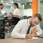 Homem sentado no trabalho, escrevendo em um papel, representando o tema FGTS e PIS são a mesma coisa.